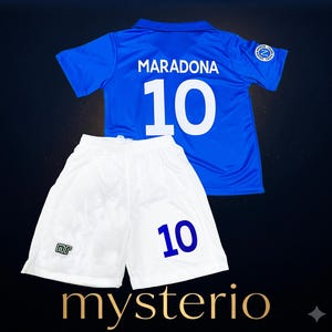 Puede incluir: Una camiseta de fútbol azul con el nombre MARADONA y el número 10 impresos en blanco en la espalda, junto con unos pantalones cortos de fútbol blancos con el número 10 en azul y un pequeño logotipo en la pierna. Incluye un escudo circular en la manga.