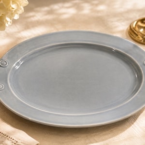 May include: An oval-shaped serving platter with a smooth, glossy light blue glaze. The platter features a raised rim and two decorative handles on opposite sides, each accented with a small circular embossed detail at the connection points.