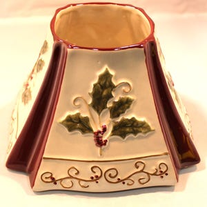 May include: A cream-colored ceramic candle topper with a flared, tapered shape. It features embossed green holly leaves and red berries on each side, accented by deep burgundy vertical trim and a delicate brown vine pattern along the base.