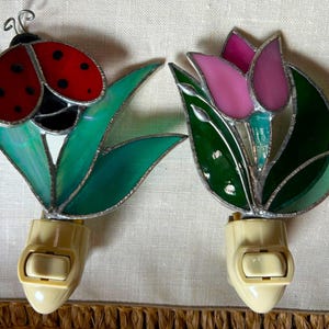 May include: Two stained glass night lights with off-white plastic plugs. One features a red ladybug with black spots on green leaves. The other displays a pink tulip flower with dark green leaves. Both have silver metal solder lines between glass pieces.