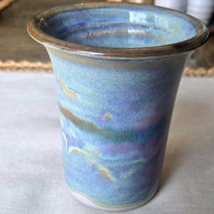May include: A ceramic tumbler with a flared rim, featuring a variegated glaze in shades of blue, purple, and hints of yellow. The surface has a smooth, glossy finish with a dark brown rim and a light-colored base, showing a hand-crafted appearance.