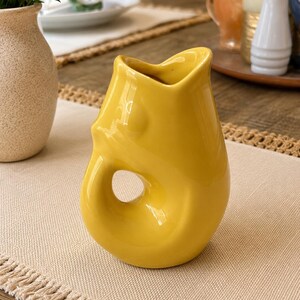 May include: A bright yellow ceramic pitcher with a smooth, glossy finish. The vessel features a distinct, organic shape with a prominent circular cutout handle integrated into the body and a wide, curved spout at the top for pouring liquids.