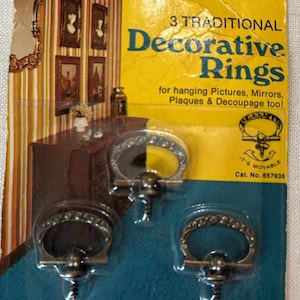 May include: A package of three metal decorative rings for hanging pictures, mirrors, and plaques. The yellow and blue packaging features the text Homecraft 3 TRADITIONAL Decorative Rings, It's Movable, and Cat. No. 657938, with Gries Reproducer Co. details.