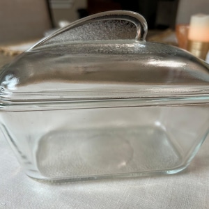 May include: A clear glass butter dish with a rectangular base and a matching lid. The lid features a smooth, curved handle integrated into the top, while the glass body has slightly tapered sides and a flat bottom for stable placement on a surface.