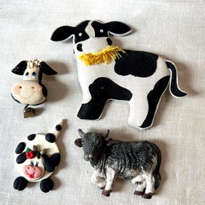May include: A collection of four cow-themed decorative items including a large fabric cow with yellow yarn, a small cow head with a metal bell, a round cow face with a red flower, and a textured gray and white cow figurine with detailed horns.