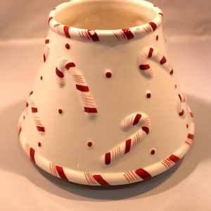 May include: A white ceramic lamp shade with a tapered, conical shape. The surface features raised, three-dimensional red and white candy cane motifs scattered across the exterior, with a matching candy cane striped pattern along the top and bottom rims.