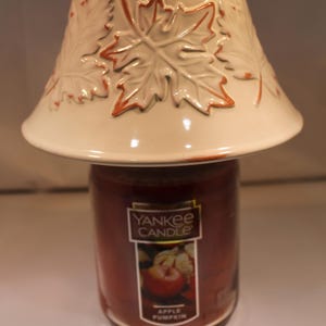 May include: A cream-colored ceramic candle topper featuring embossed maple leaf designs with orange-brown edges, resting on a glass jar of Yankee Candle Apple Pumpkin scented candle with a label showing an apple and pumpkin graphic.