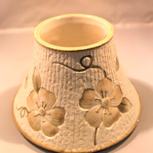May include: A conical ceramic lampshade featuring a textured, off-white surface with embossed, light brown floral motifs and vine details. The shade has a smooth, rounded top opening and a wider base, both finished with a thin, light brown trim.