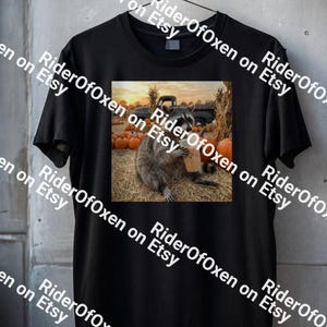 May include: A black short-sleeved t-shirt featuring a rectangular graphic print of a raccoon sitting on hay, holding a cup, with pumpkins and a truck in the background. The text RiderOfOxen on Etsy is repeated across the image as a watermark.
