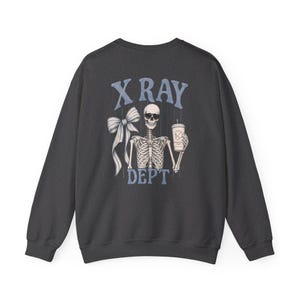 May include: A charcoal gray crewneck sweatshirt featuring a graphic on the back of a skeleton wearing sunglasses, holding a coffee cup, and positioned next to a large bow. The text X RAY DEPT is printed in light blue above and below the skeleton.