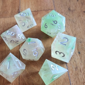May include: A set of seven polyhedral dice made from translucent, pale green resin with a marbled effect. Each die features engraved numbers filled with silver-colored ink, including a twenty-sided die, a twelve-sided die, and various others.