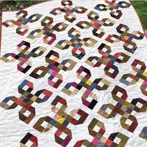 Puede incluir: Una colcha con fondo blanco y un patrón repetitivo de estrellas de cuatro puntas formadas por tiras de tela de varios colores. La colcha tiene un borde rojo fino y costuras de acolchado onduladas visibles en toda la superficie.