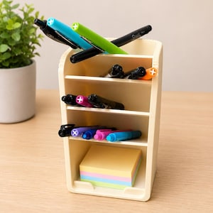 May include: A cream-colored plastic desk organizer with four horizontal shelves and a vertical compartment. The organizer holds various colored pens and markers, including black, blue, green, and pink, along with a stack of multi-colored sticky notes.