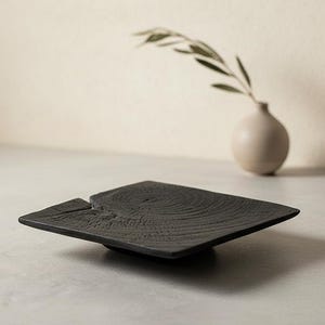 May include: A square, matte black wooden tray featuring a prominent natural wood grain texture and a visible crack detail on one side. The tray has a low profile and a flat surface, showcasing the organic patterns of the wood grain throughout.