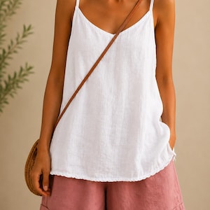 May include: A white sleeveless camisole top with thin shoulder straps and a V-neckline, paired with dusty rose pink shorts featuring cuffed hems. A round woven straw bag with a brown leather strap hangs across the torso of the garment.