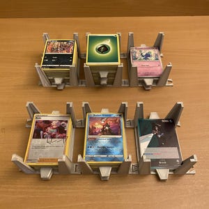May include: Two grey plastic card organizer racks holding six stacks of trading cards. The top row features a dark card, a green energy card, and a pink card. The bottom row displays a trainer card, a blue Radiant Greninja card, and a Brook card.