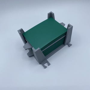 May include: A gray 3D-printed plastic card holder containing a stack of dark green rectangular cards. The holder features four vertical corner supports and small protruding feet at the base, designed to securely store and organize a deck of cards.
