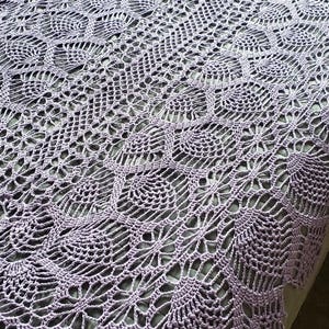 Puede incluir: Una manta de ganchillo lavanda hecha a mano con un diseño intrincado y calado. La manta presenta un patrón repetitivo de formas florales y geométricas, creando una superficie texturizada. La manta está sobre una cama, mostrando su tamaño y detalle.