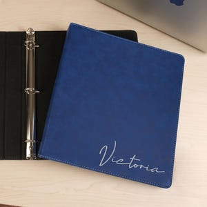 May include: A blue three-ring binder with a textured finish and the name Victoria written in a white script font on the front cover. The binder is open, revealing a black interior with a metal ring mechanism and a pocket on the left side.