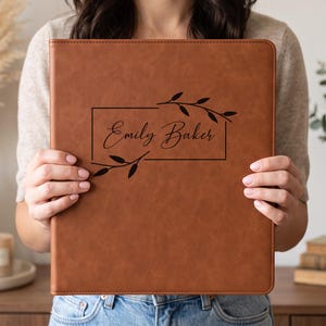 May include: A brown leather-like portfolio folder featuring a rectangular frame design with black text that reads Emily Baker, accented by two black leafy branch illustrations extending from the corners of the frame on the front cover.