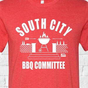 May include: Red t-shirt with a white graphic design that says "South City BBQ Committee". The graphic features a grill with flames, a brick wall, and a bottle opener.