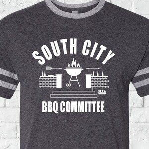 May include: A dark heather grey t-shirt with white stripes on the sleeves. The shirt has a white graphic of a grill with flames, beer bottles, and the text "South City BBQ Committee".