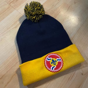 以下が含まれることがあります： ネイビーブルーのニット帽。黄色のつばと黄と黒のポンポンが付いています。 赤、白、黄色のフルール・ド・リスのデザインが施された円形エンブレムが特徴です。