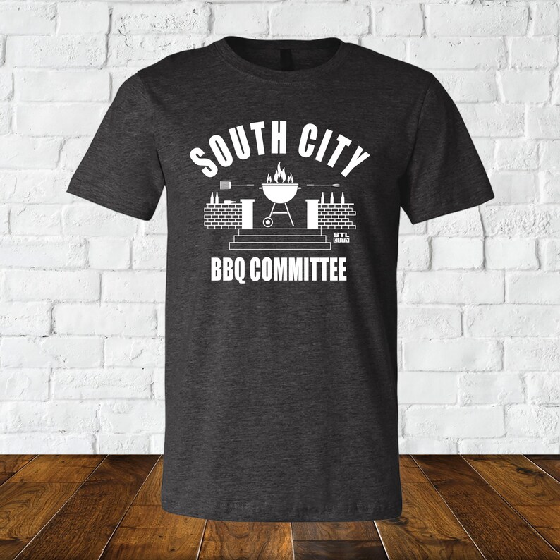 May include: A dark heather grey t-shirt with a white graphic design. The graphic features a grill with flames, beer bottles, and the text "South City BBQ Committee".