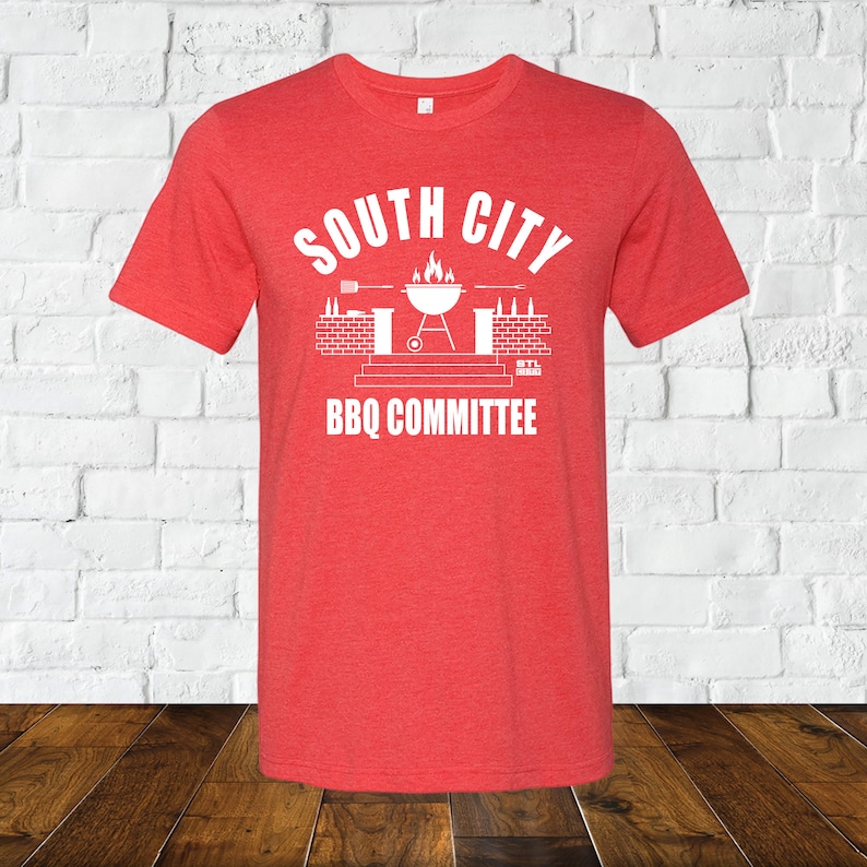 May include: Red t-shirt with a white graphic design that says "South City BBQ Committee". The graphic features a grill with flames, a brick wall, and a bottle opener.