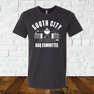 May include: A dark gray t-shirt with a white graphic design. The graphic features a grill with flames, a brick wall, and the text "South City BBQ Committee".