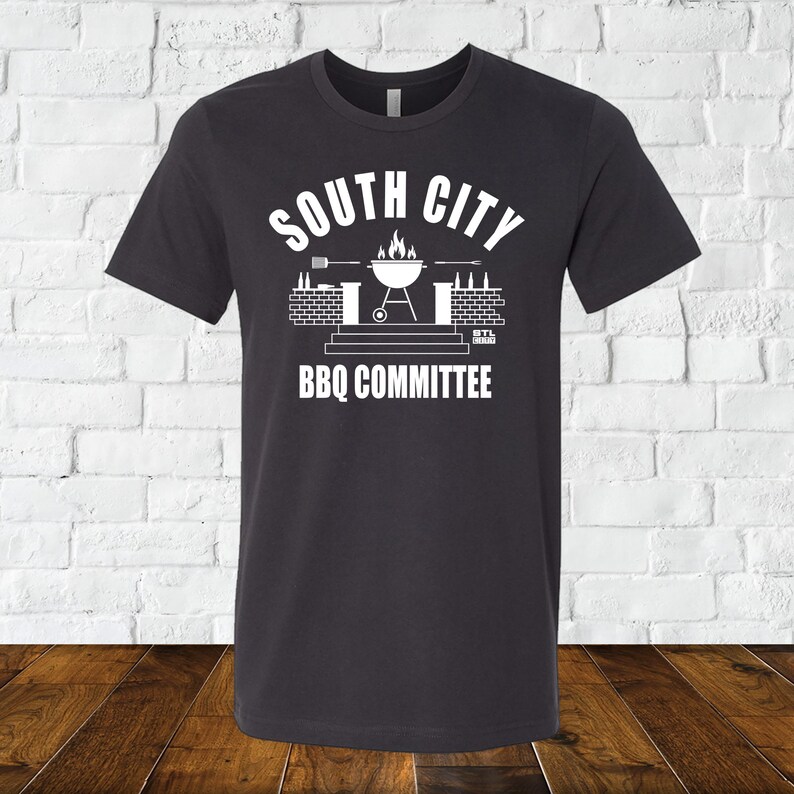 May include: A dark gray t-shirt with a white graphic design. The graphic features a grill with flames, a brick wall, and the text "South City BBQ Committee".