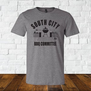 May include: A heather gray t-shirt with a black graphic design. The graphic features a grill with flames, a brick wall, and the text "South City BBQ Committee".