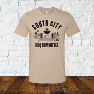 May include: Tan t-shirt with a black graphic of a grill, brick wall, and the text "South City BBQ Committee".