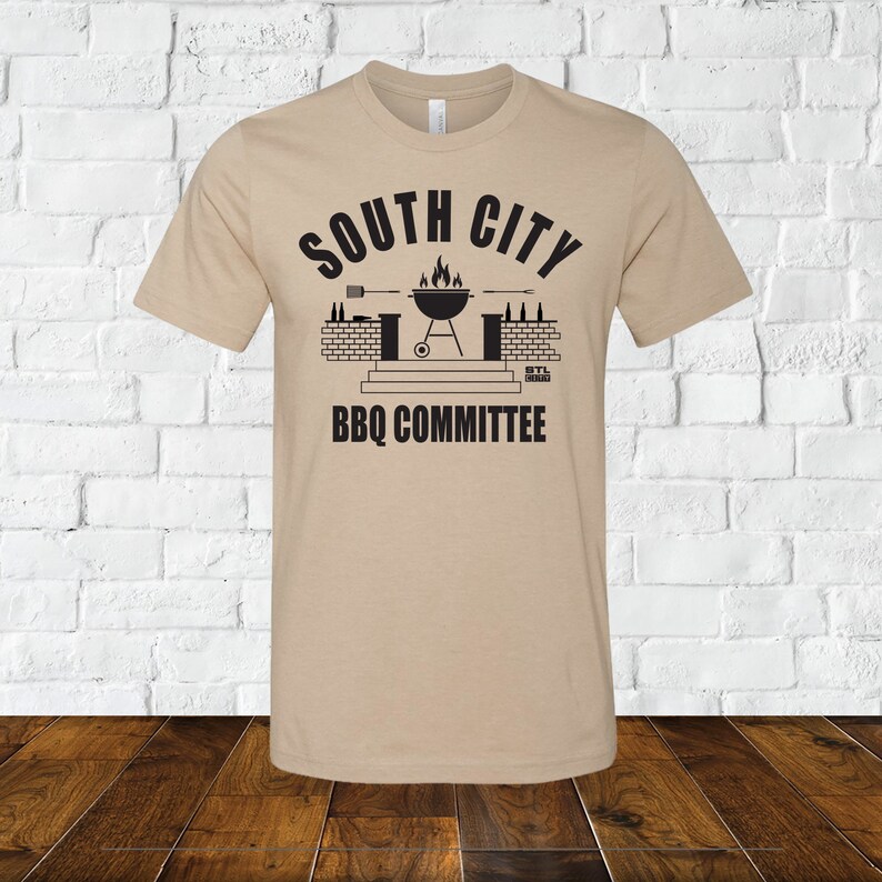 May include: Tan t-shirt with a black graphic of a grill, brick wall, and the text "South City BBQ Committee".