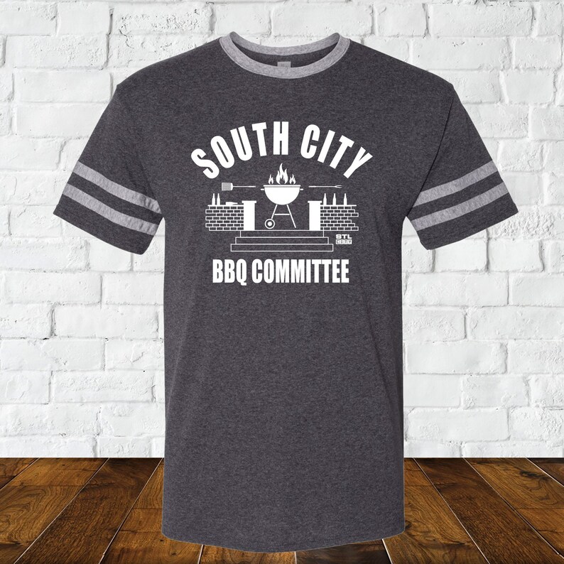 May include: A dark heather grey t-shirt with white stripes on the sleeves. The shirt has a white graphic of a grill with flames, beer bottles, and the text "South City BBQ Committee".