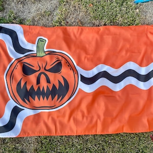May include: An orange flag with a black and white wavy design. A large, cartoon jack-o'-lantern face is centered on the flag.