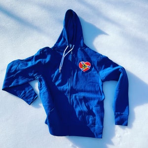 Puede incluir: Una sudadera con capucha azul real con un parche en forma de corazón que muestra la bandera de la ciudad de St. Louis.