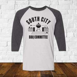 May include: A white and gray baseball tee shirt with a black graphic of a grill, brick wall, and the text "South City BBQ Committee".
