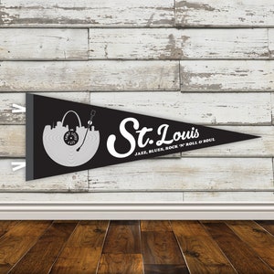 May include: Black pennant flag with a white silhouette of the St. Louis skyline and a record player. The text "St. Louis" is written in a white cursive font with the words "Jazz, Blues, Rock 'n' Roll & Soul" underneath.