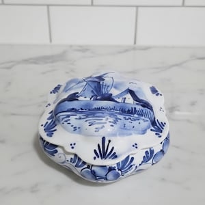 May include: A white ceramic trinket box with a scalloped edge and blue hand-painted details. The lid features a detailed illustration of a windmill in a landscape, while the base is decorated with blue floral and leaf patterns around the perimeter.