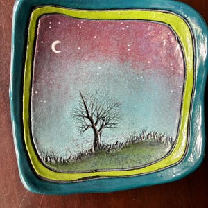 Pode incluir: Um prato quadrado de cerâmica com bordas arredondadas apresentando uma cena noturna pintada à mão. O centro retrata uma árvore nua numa colina relvada sob um céu roxo e azul com uma lua crescente branca e estrelas. Borda verde-limão e azul-petróleo.