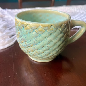 May include: A ceramic mug featuring a textured, raised scale pattern across its exterior. The mug is finished in a glossy glaze with blended shades of light green, turquoise, and hints of golden-brown along the edges of the scales and handle.