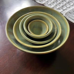 May include: A set of four nested ceramic bowls with a glossy, variegated sage green and olive glaze. The bowls are arranged in a concentric stack, with each bowl decreasing in size from the outermost to the innermost, featuring smooth, rounded rims.