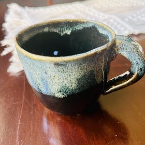 May include: A ceramic mug featuring a two-tone glaze with a dark brown base and a textured, mottled light blue and cream upper section. The mug has a wide, rounded body and a thick, curved handle that matches the blue and cream color scheme.
