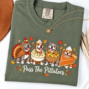 May include: An olive green t-shirt featuring a graphic of four dogs dressed as Thanksgiving food: a turkey, mashed potatoes, stuffing, and pumpkin pie. The text below reads Pass The Pittatoes. A gold chain necklace rests on the collar.