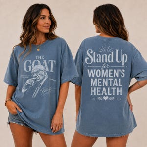 以下が含まれることがあります： 前面と背面にグラフィックがプリントされた青い半袖Tシャツ。前面には男性の肖像画と「THE GOAT GREATEST OF ALL TIME」の文字。背面には「Stand Up for WOMEN'S MENTAL HEALTH」という文字とハートのアイコンがデザインされています。
