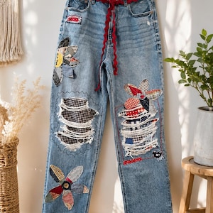 May include: Light blue denim jeans featuring a high waist with a red rickrack trim belt. The pants are decorated with various fabric flower patches, distressed areas with plaid and polka dot fabric backing, and red rickrack trim at the hem.
