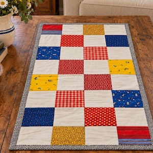 May include: A rectangular patchwork table runner featuring a grid of alternating solid cream squares and various patterned fabric squares in red, blue, and yellow. The runner is finished with a thin grey and white speckled border trim.