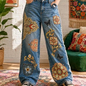 Puede incluir: Pantalones vaqueros azules de corte ancho con costuras laterales deshilachadas y varios parches bordados coloridos, incluyendo motivos florales, patrones geométricos complejos, mariposas negras y un pequeño parche de yin-yang en el bolsillo.
