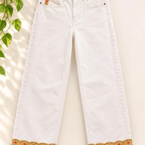 Puede incluir: Pantalones vaqueros de color blanco roto con cintura alta, pernera ancha y cierre de botones. El bajo de cada pernera presenta un borde festoneado y una banda decorativa de flores rojas bordadas sobre un fondo amarillo mostaza.